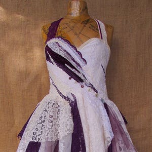 Puede incluir: Un vestido de encaje blanco y morado de varias capas con una falda de tul. El vestido tiene un corpi&ntilde;o ajustado y una falda fluida. El vestido est&aacute; decorado con botones y encaje.
