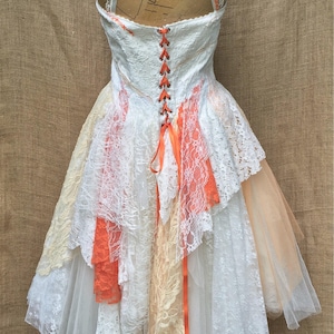 May include: A white lace wedding dress with a layered skirt and a peach-colored ribbon tied at the back. The dress is on a mannequin and has a lace-up back.