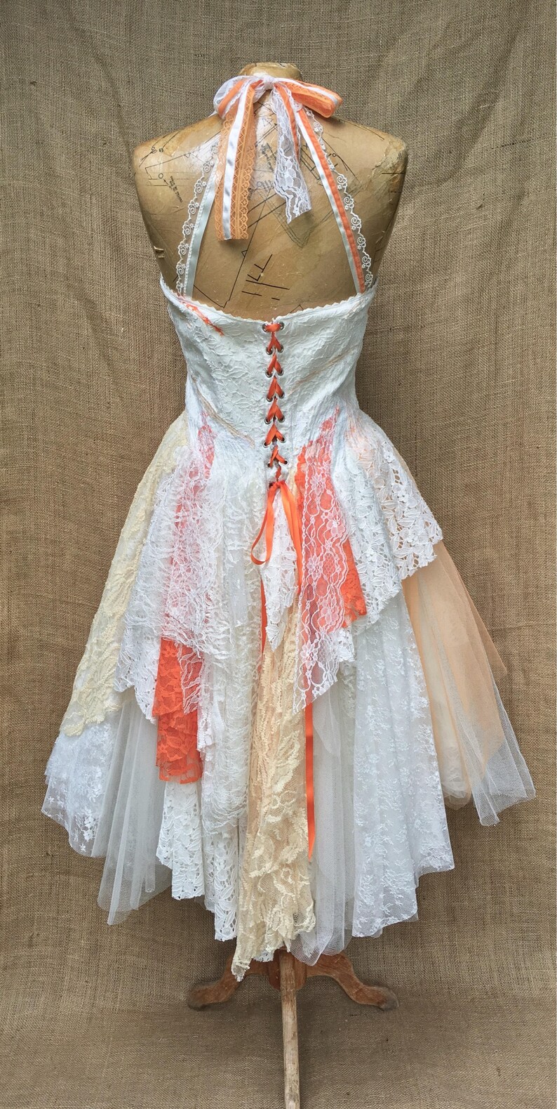 May include: A white lace wedding dress with a layered skirt and a peach-colored ribbon tied at the back. The dress is on a mannequin and has a lace-up back.