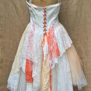 Puede incluir: Un vestido de novia de encaje blanco con una falda de capas y una cinta de color melocot&oacute;n atada en la espalda. El vestido est&aacute; en un maniqu&iacute; y tiene una espalda con cordones.