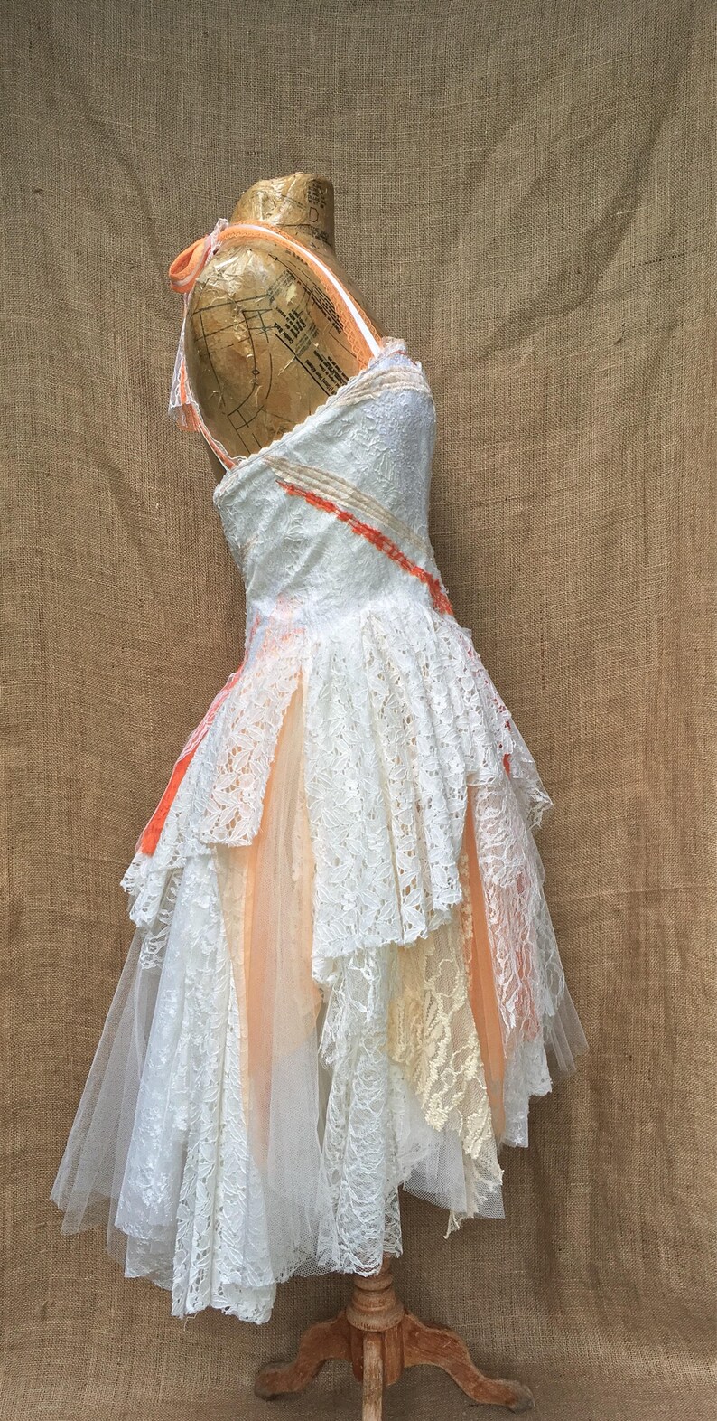 May include: A white lace dress with a peach tulle overlay and a peach ribbon detail. The dress is on a dress form and has a layered skirt.