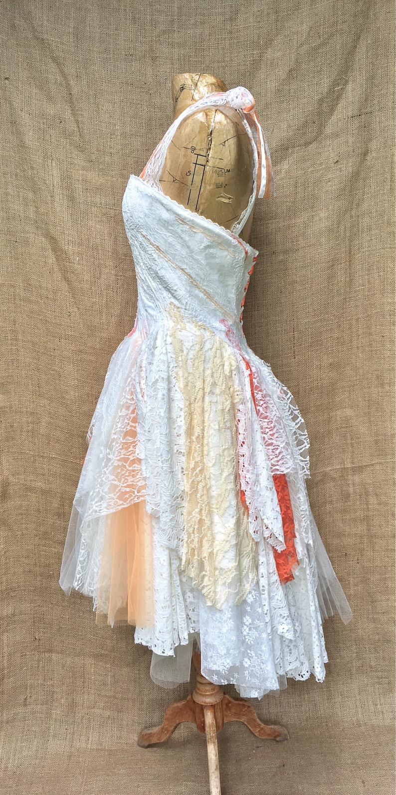 Puede incluir: Un vestido de encaje blanco con capas de tul y encaje en tonos melocot&oacute;n, crema y naranja. El vestido est&aacute; en un maniqu&iacute; y tiene un cierre de bot&oacute;n en la espalda.
