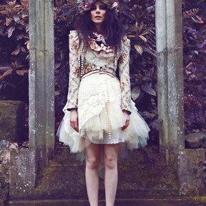 May include: A woman wearing a white dress with a floral print and a tulle overlay. She is wearing a flower crown and standing in front of a stone wall.