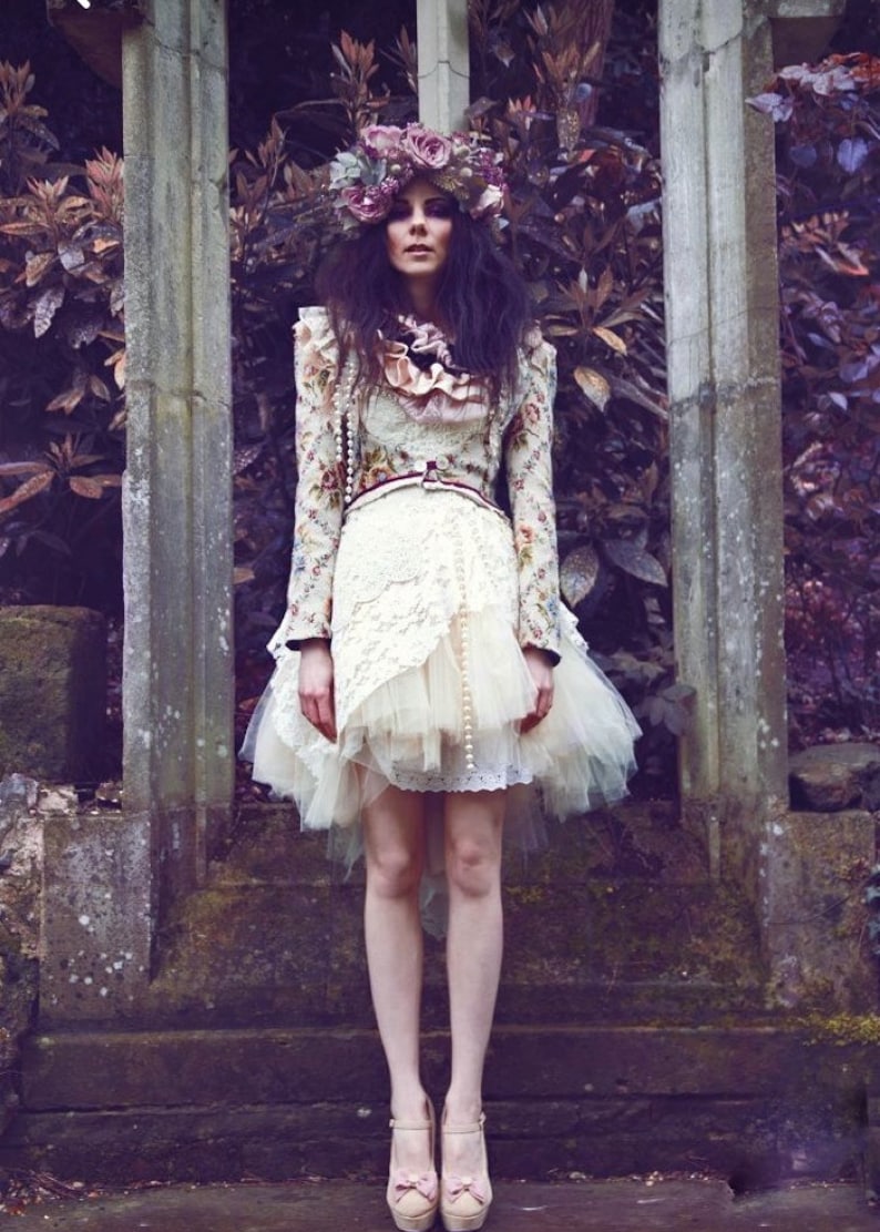 May include: A woman wearing a white dress with a floral print and a tulle overlay. She is wearing a flower crown and standing in front of a stone wall.