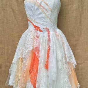 Puede incluir: Un vestido blanco de encaje con escote halter y falda de capas. La falda tiene un degradado de color melocot&oacute;n y naranja. El vestido est&aacute; en un maniqu&iacute;.