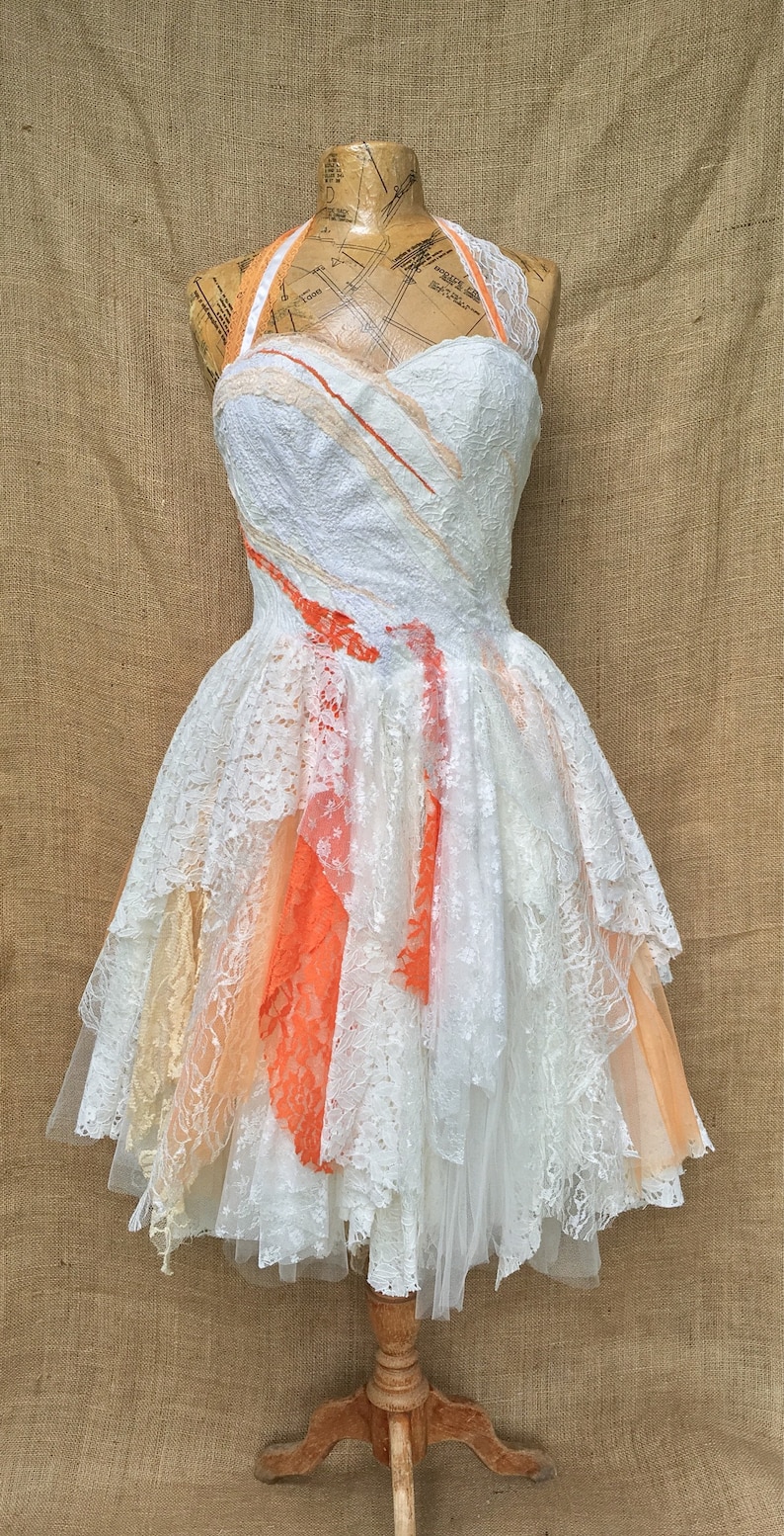 Puede incluir: Un vestido blanco de encaje con escote halter y falda de capas. La falda tiene un degradado de color melocot&oacute;n y naranja. El vestido est&aacute; en un maniqu&iacute;.