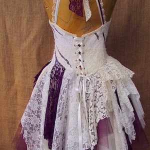 May include: A white and purple layered lace dress with a white satin ribbon tied at the back. The dress has a fitted bodice with a lace-up back and a flowing skirt.