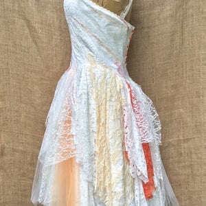 May include: A white lace dress with layers of tulle and lace in shades of peach, cream, and orange. The dress is on a dress form and has a button closure in the back.