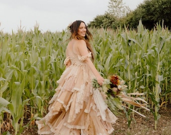 May include: A woman in a tiered, off-the-shoulder, beige lace dress walks through a cornfield, holding a bouquet of flowers. The dress has multiple layers of lace and a flowing skirt. The background features tall green corn stalks.