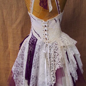 Puede incluir: Un vestido de encaje blanco y morado con espalda de cors&eacute; y falda de capas. El vestido est&aacute; atado con una cinta blanca en la espalda.