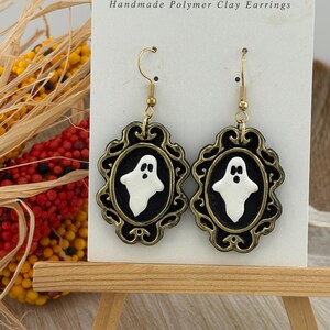 May include: A pair of dangle earrings featuring a white ghost figure centered on a black oval background, encased in an ornate, antique gold-colored frame. The earrings hang from gold-toned metal hooks on a white display card labeled Bejeweled by Chris.
