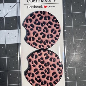 May include: Two car coasters with a pink leopard print design. The coasters are round with a small notch on one side. The packaging reads "Car Coasters Handmade with love."