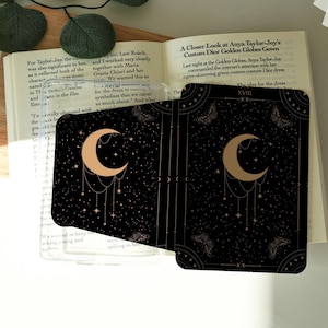 May include: Two tarot cards with a black background featuring a gold crescent moon, stars, and decorative elements. The cards are placed on top of an open book. The text "XVIII" is visible on one card.