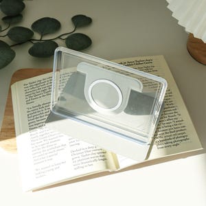 May include: A clear, rectangular phone case with a silver ring holder is placed over an open book. The book's pages are visible, and the case is designed to hold a phone for hands-free use. The background includes greenery.
