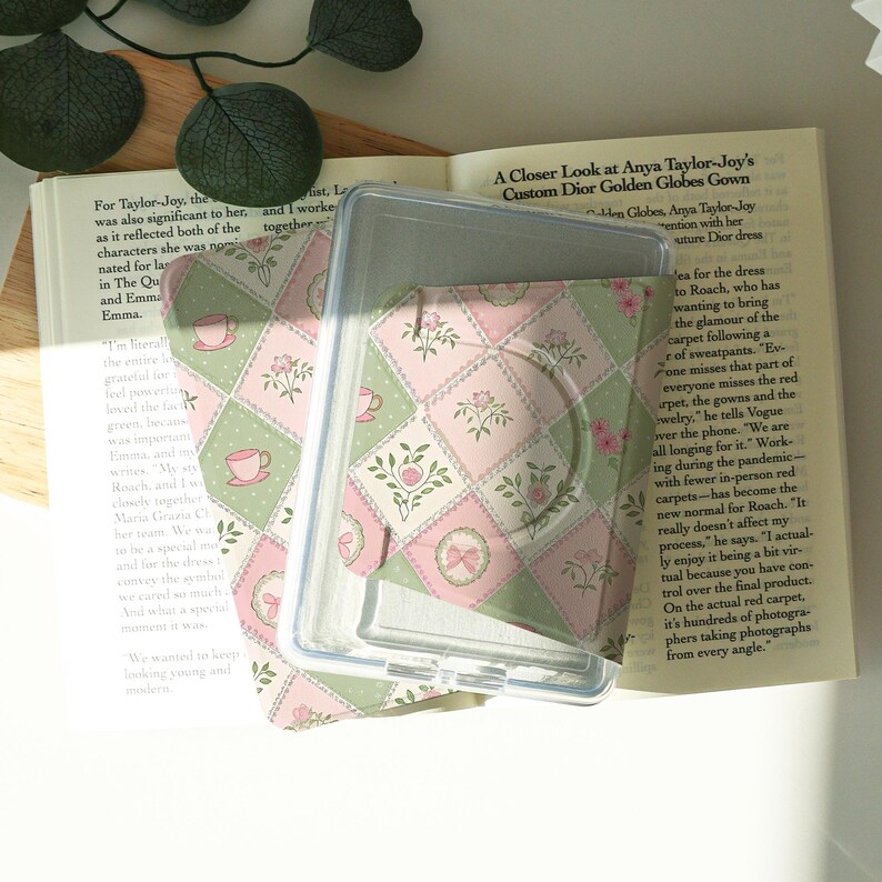 May include: A clear plastic phone case with a pink and green floral patterned insert. The insert features a repeating design of teacups, flowers, and bows. The case is open, revealing the patterned insert.