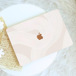 May include: A laptop case with a beige background and a white, wavy pattern. The Apple logo is visible in the center. The case is on a white table with a piece of paper and a flower.