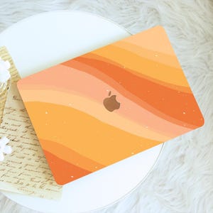 May include: A laptop case with a warm color palette of orange and peach tones, featuring a wavy design. The Apple logo is visible in the center. The case is on a white surface with a vintage-style paper and white flower.