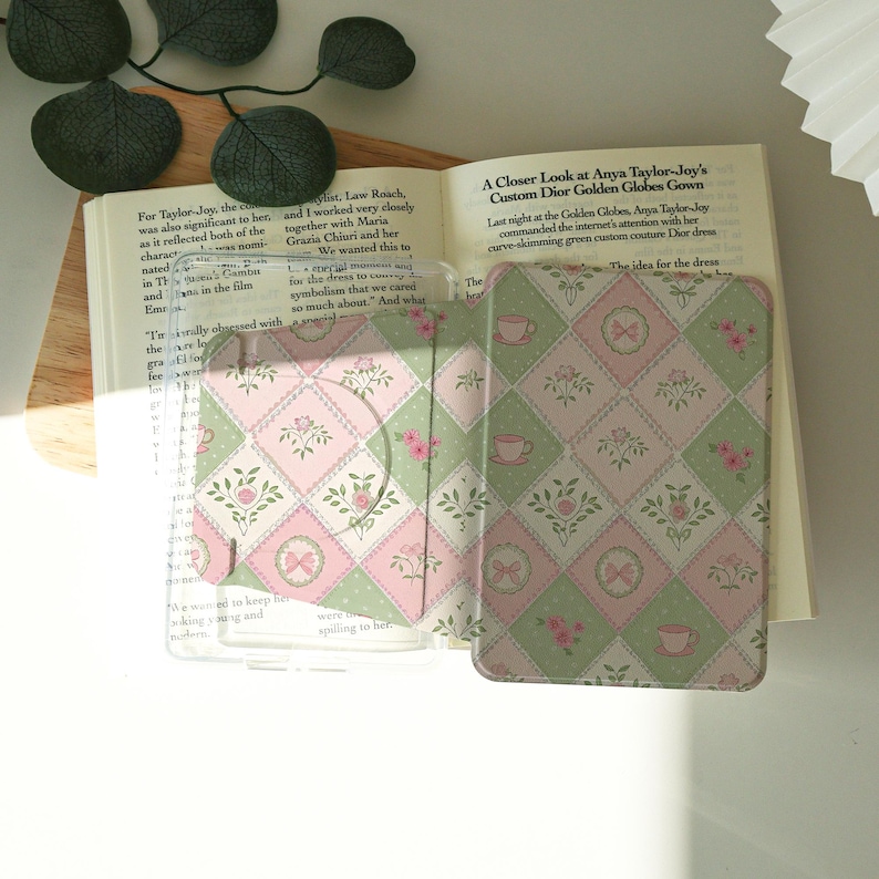 May include: A clear, open card holder with a pink and green floral pattern, featuring teacups and bows. The card holder is open, revealing the interior. The background includes an open book.