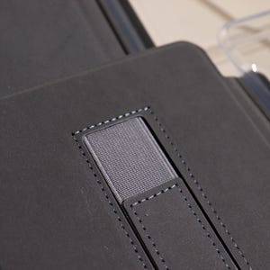 May include: Close-up of a dark gray leather case with a stitched elastic band and a small leather tab. The case appears to be for a tablet or similar device, with a minimalist design.