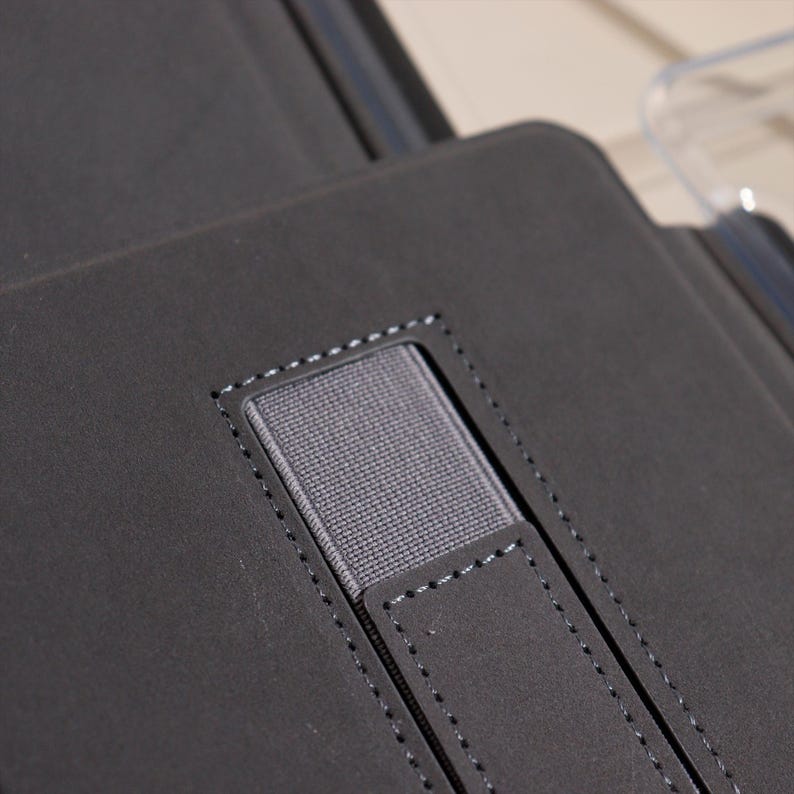 May include: Close-up of a dark gray leather case with a stitched elastic band and a small leather tab. The case appears to be for a tablet or similar device, with a minimalist design.