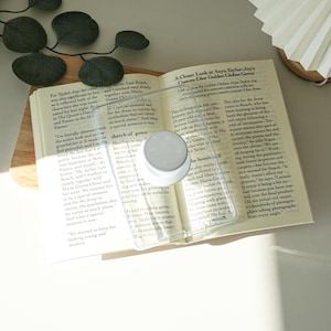 May include: A clear, rectangular phone case with a white circular pop-up grip is placed on an open book. The book is on a wooden tray, and the pages display printed text. The scene is lit with natural light.