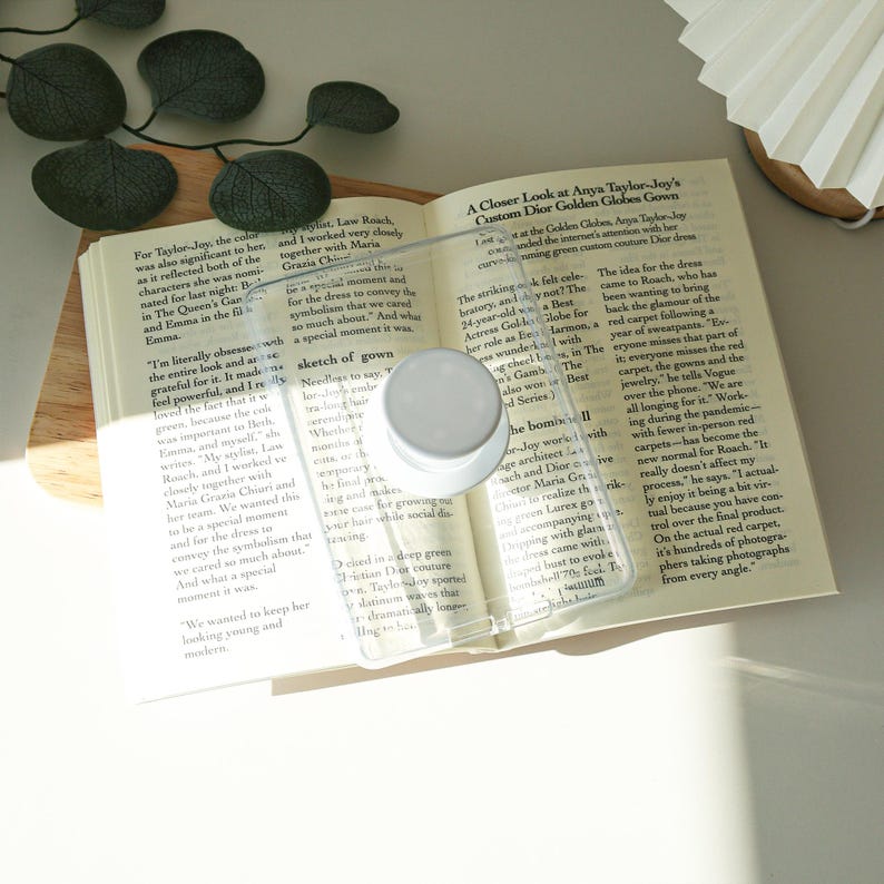 May include: A clear, rectangular phone case with a white circular pop-up grip is placed on an open book. The book is on a wooden tray, and the pages display printed text. The scene is lit with natural light.