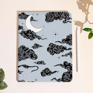 May include: A notebook with a light blue cover featuring a celestial design. The design includes black swirling clouds, a white crescent moon, stars, and a stylus. The notebook is a rectangular shape.