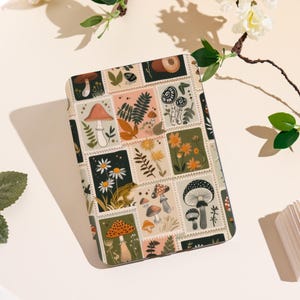 Może przedstawiać: Prostokątne etui z fantazyjnym wzorem grzybów i kwiatów. Etui posiada kolaż kwadratów przypominających znaczki pocztowe z ilustracjami grzybów, stokrotek, paproci i innych elementów botanicznych.