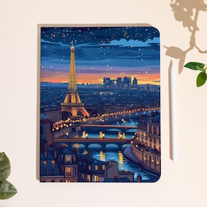 Könnte beinhalten: Ein Notizbuch mit einer lebendigen Illustration des Eiffelturms in Paris bei Dämmerung. Das Kunstwerk zeigt den beleuchteten Turm, Stadtlichter und einen Fluss, vor einem Abendhimmel mit Sternen. Ein weißer Stift befindet sich rechts.