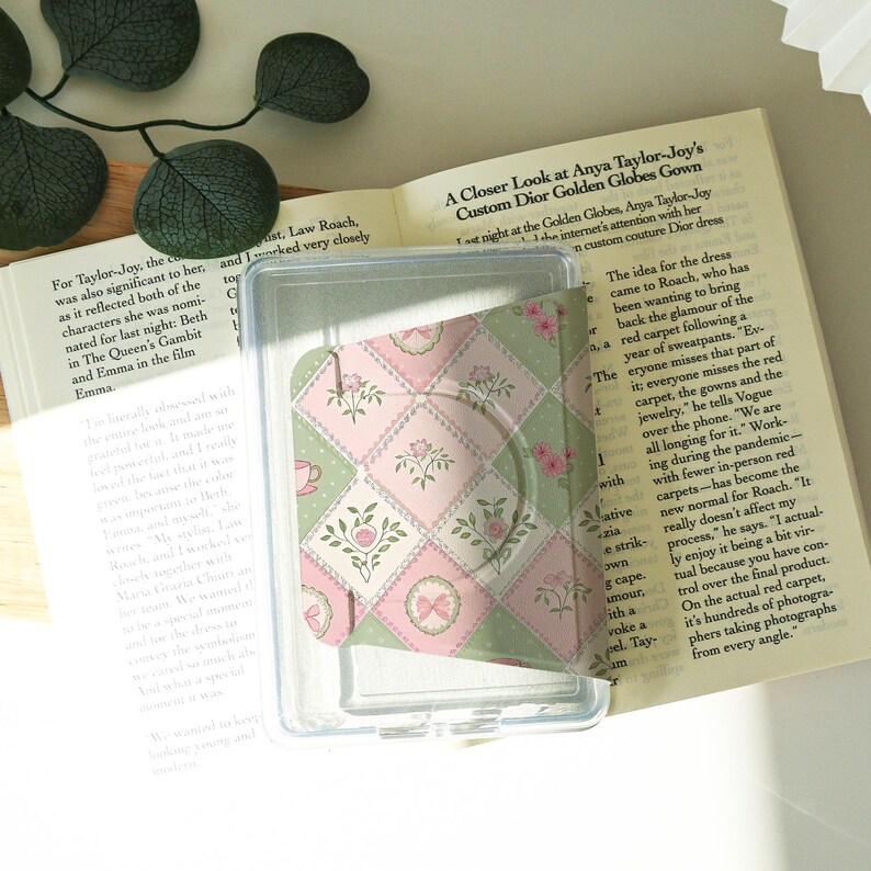 May include: A clear plastic phone grip with a pink, green, and white floral pattern. The grip is in a clear plastic case, resting on an open book. The pattern includes teacups, flowers, and bows.