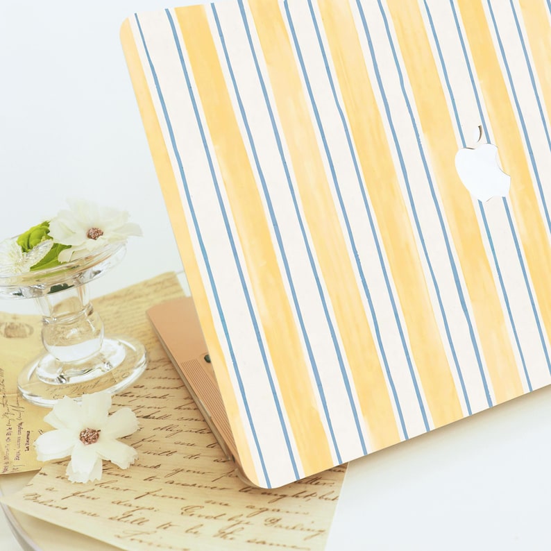 May include: Laptop with a cover featuring a pattern of yellow and blue vertical stripes on a white background. The Apple logo is visible on the cover. The laptop is partially open, revealing a rose gold interior. A glass dish with white flowers is in the foreground.