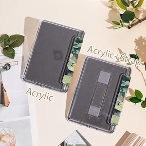 May include: Two clear acrylic tablet cases with a gray interior. One case has a strap with a green and blue floral design. The text "Acrylic" and "Acrylic+Strap" are visible. The cases are displayed on a light beige surface.