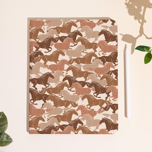 Può includere: Un quaderno con un motivo ripetuto di cavalli in corsa nei toni del marrone, beige e crema. Il quaderno è rettangolare con angoli arrotondati. Uno stilo bianco è accanto al quaderno.
