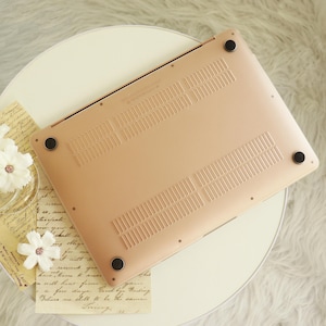 May include: A rose gold laptop computer, viewed from the bottom, with black rubber feet. The underside has ventilation slots and a smooth, metallic finish. The laptop is positioned on a white surface with decorative elements.