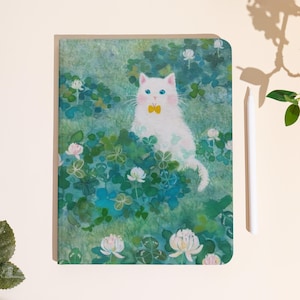Puede incluir: Un cuaderno con una ilustración caprichosa de un gato blanco con ojos azules y una pajarita amarilla, rodeado de tréboles verdes y flores blancas. La portada tiene un estilo acuarela con una estética suave y soñadora.