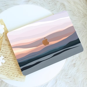 May include: A laptop case featuring a sunset-inspired design with shades of pink, orange, and blue. The Apple logo is visible. The case is on a white surface with a piece of paper.
