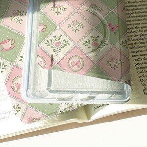 May include: Clear phone case with a pastel pink and green floral pattern. The design features a patchwork of squares with floral and bow motifs. The case is partially covering a book.