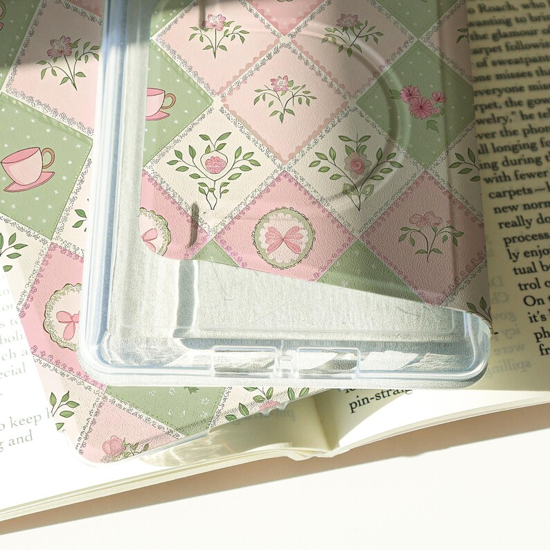 May include: Clear phone case with a pastel pink and green floral pattern. The design features a patchwork of squares with floral and bow motifs. The case is partially covering a book.