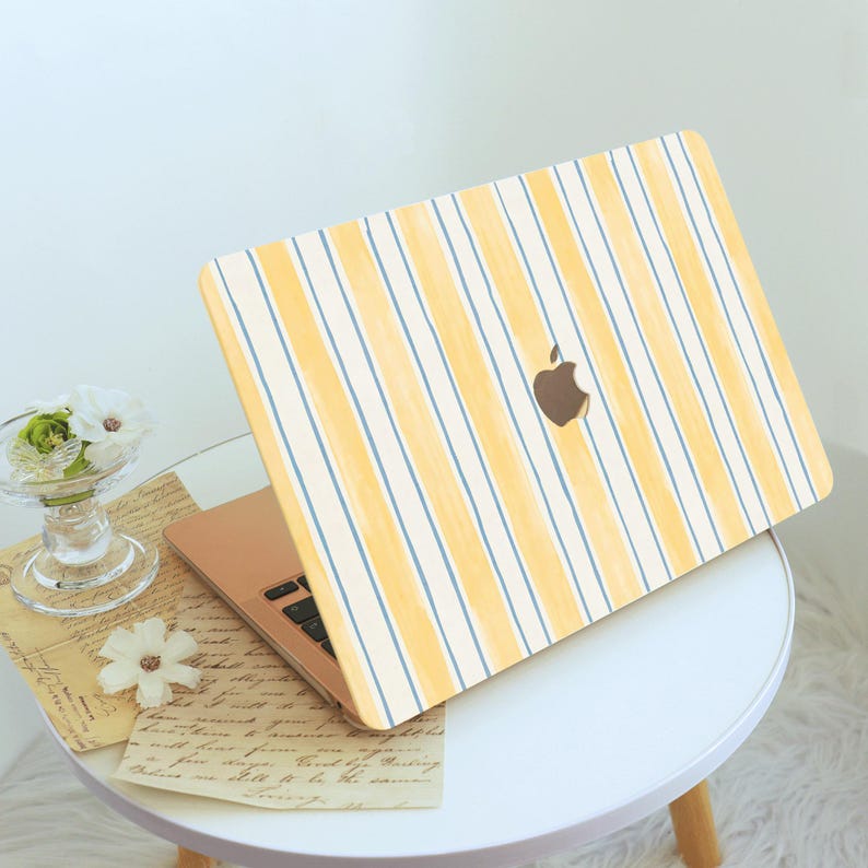 May include: A laptop with a yellow and blue striped cover, featuring the Apple logo. The laptop is open on a small white table, with a glass vase of flowers and vintage papers nearby. The cover design has vertical yellow stripes with thin blue lines.