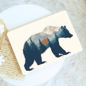 Könnte beinhalten: Eine cremefarbene Laptop-Hülle mit einer Bärensilhouette und einem Berg- und Waldlandschaftsdesign. Der Körper des Bären integriert eine blau-weiße Bergszene mit einem orangefarbenen Apple-Logo in der Mitte.