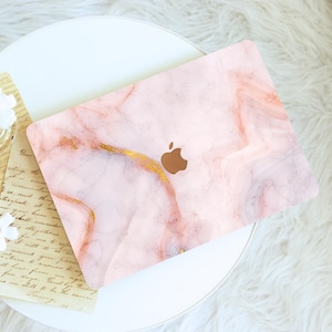 May include: A laptop with a pink marble design and gold accents. The laptop has a brown Apple logo on the top. The laptop is on a white table with a white fluffy rug in the background.