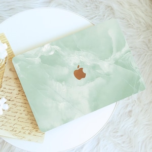 May include: A laptop featuring a light green marble effect and a copper-coloured Apple logo. The laptop sits on a white surface, with a piece of paper with handwritten text visible. The design is clean and modern, with a focus on aesthetics.