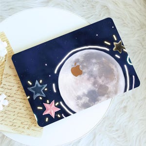 May include: A dark blue laptop skin featuring a large, detailed full moon graphic with a gold Apple logo cutout. The design includes a white hand-drawn circular outline around the moon, along with small blue and pink stars and white light accents.