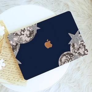 May include: A navy blue laptop case featuring a design with silver disco balls and glittery stars. The case has a gold Apple logo in the center. The design is on a white table with a fluffy white rug.