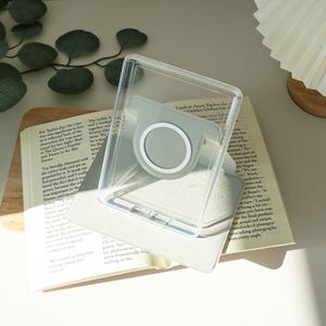 May include: A clear phone case with a gray magnetic ring holder, resting on an open book. The case is transparent, allowing the book's pages to be visible. The book has text and is open to a page with text. The background is neutral.