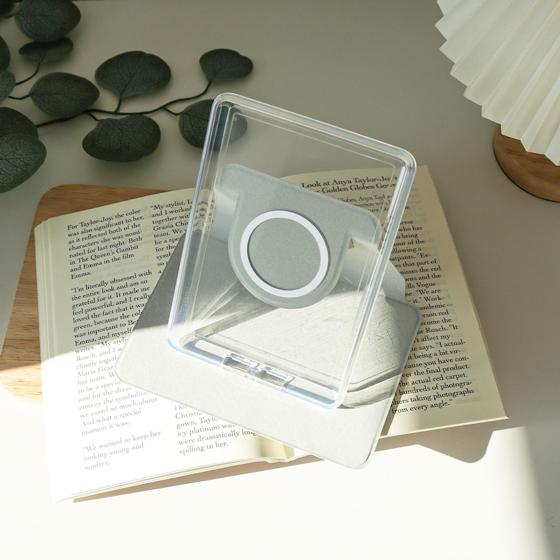 May include: A clear phone case with a gray magnetic ring holder, resting on an open book. The case is transparent, allowing the book's pages to be visible. The book has text and is open to a page with text. The background is neutral.
