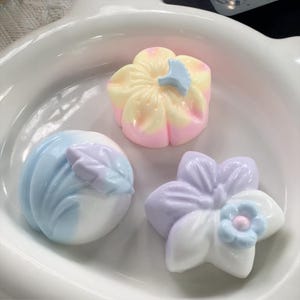 Op de afbeelding: Drie pastelkleurige, bloemvormige decoraties in een witte kom. Eén is geel en roze, een andere is blauw en wit, en de derde is paars en wit met een blauw centrum.