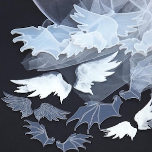 May include: An assortment of translucent, wing-shaped embellishments in various sizes and styles. Some wings are detailed with feathery textures, while others have a smooth, solid appearance. The wings are arranged on a sheer, white fabric against a dark background.