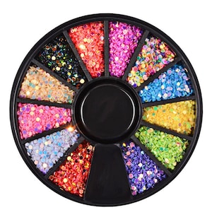 Puede incluir: Un recipiente negro circular dividido en doce secciones, cada una llena de lentejuelas iridiscentes de colores. Las lentejuelas son de varios colores, incluyendo rojo, rosa, azul, verde y dorado, creando una exhibición vibrante y llamativa. Ideal para arte de uñas o manualidades.