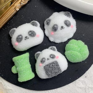 May include: Close-up of several white, panda-shaped candies with black ears and eyes, and pink cheeks. Also includes green, leaf-shaped and bamboo-shaped candies. The candies are on a black surface.